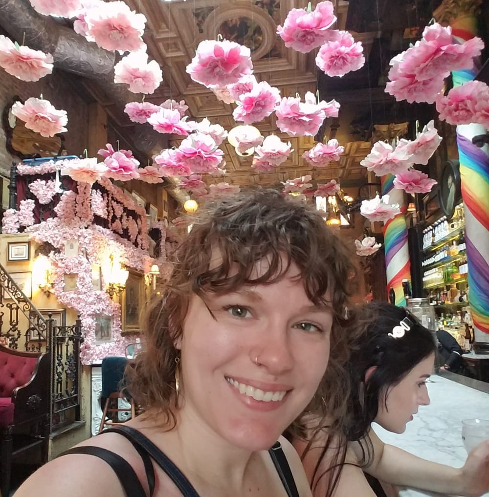 Woman with messy curly hair and banks smiling at camera in a vintage-looking restaurant with a bar and decorated with pink f