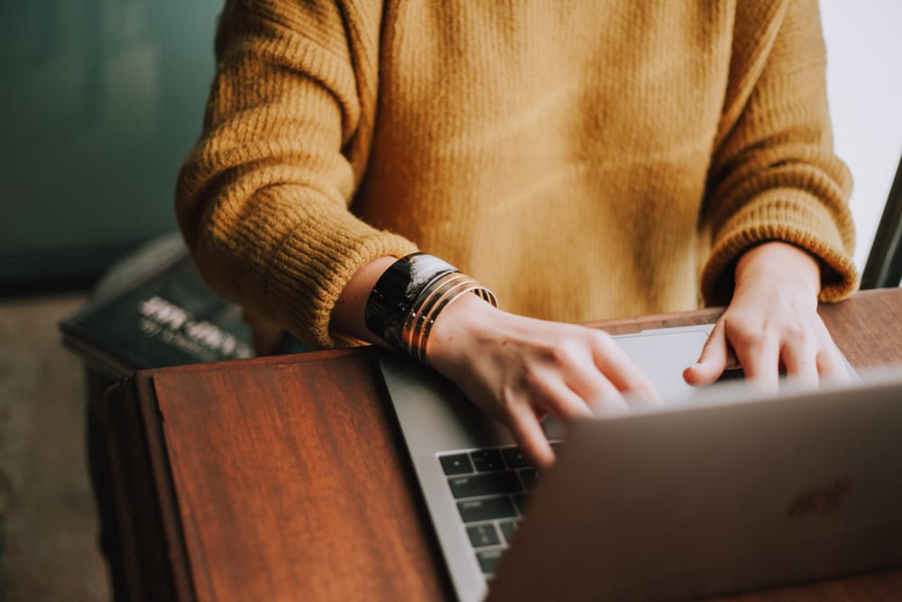 Neck-down shot of person in golden brown sweater with gold bracelet typing on laptop at wooden table.