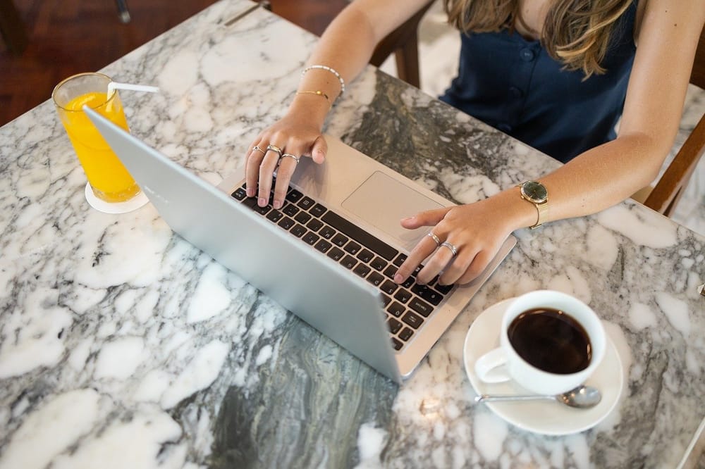 Downward shot of woman working a full-time remote job on her computer at a marble table with a coffee cup and beverage.