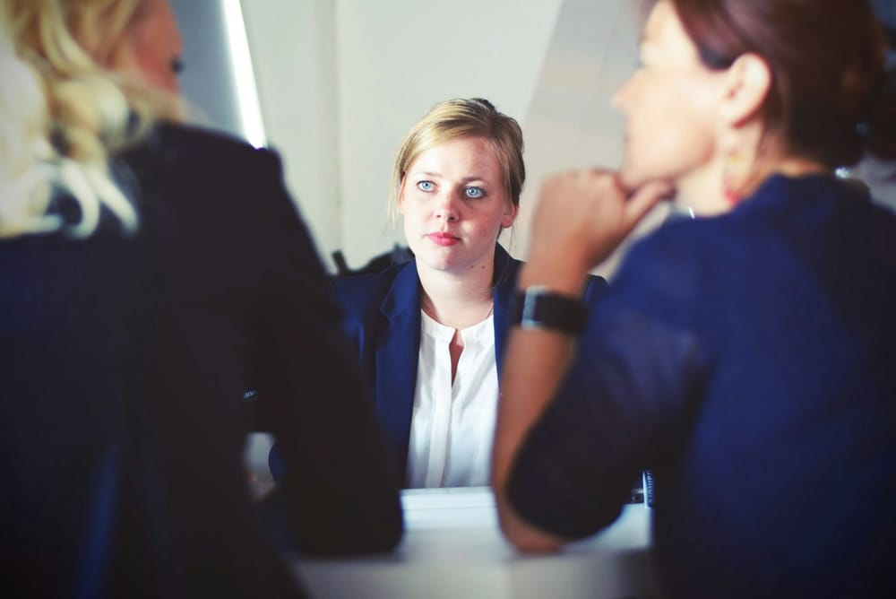 Two women interviewers in suits with their backs to the camera face a young job candidate with blones hair and blue eyes.