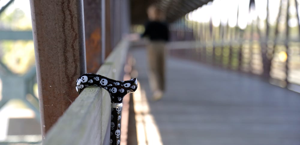 Close up of black walking cane with white skulls leaning on a bidge with person walking away in background.