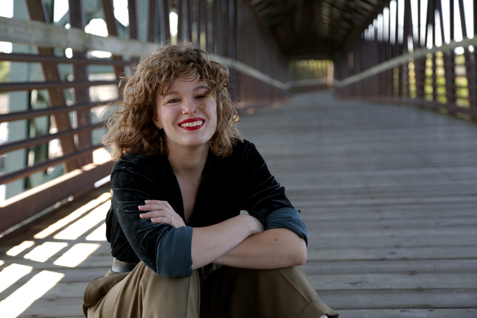 White woman with blonde hair and red lipstick sits on bridge and smiles at the camera