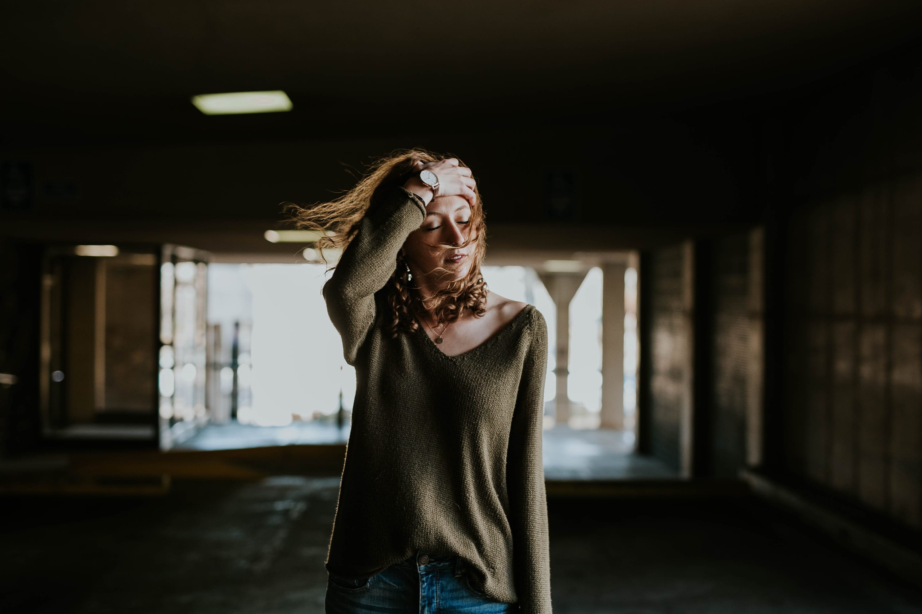 Woman standing in dark building with her eyes closed, brushing her hair out of her face like she's tired.