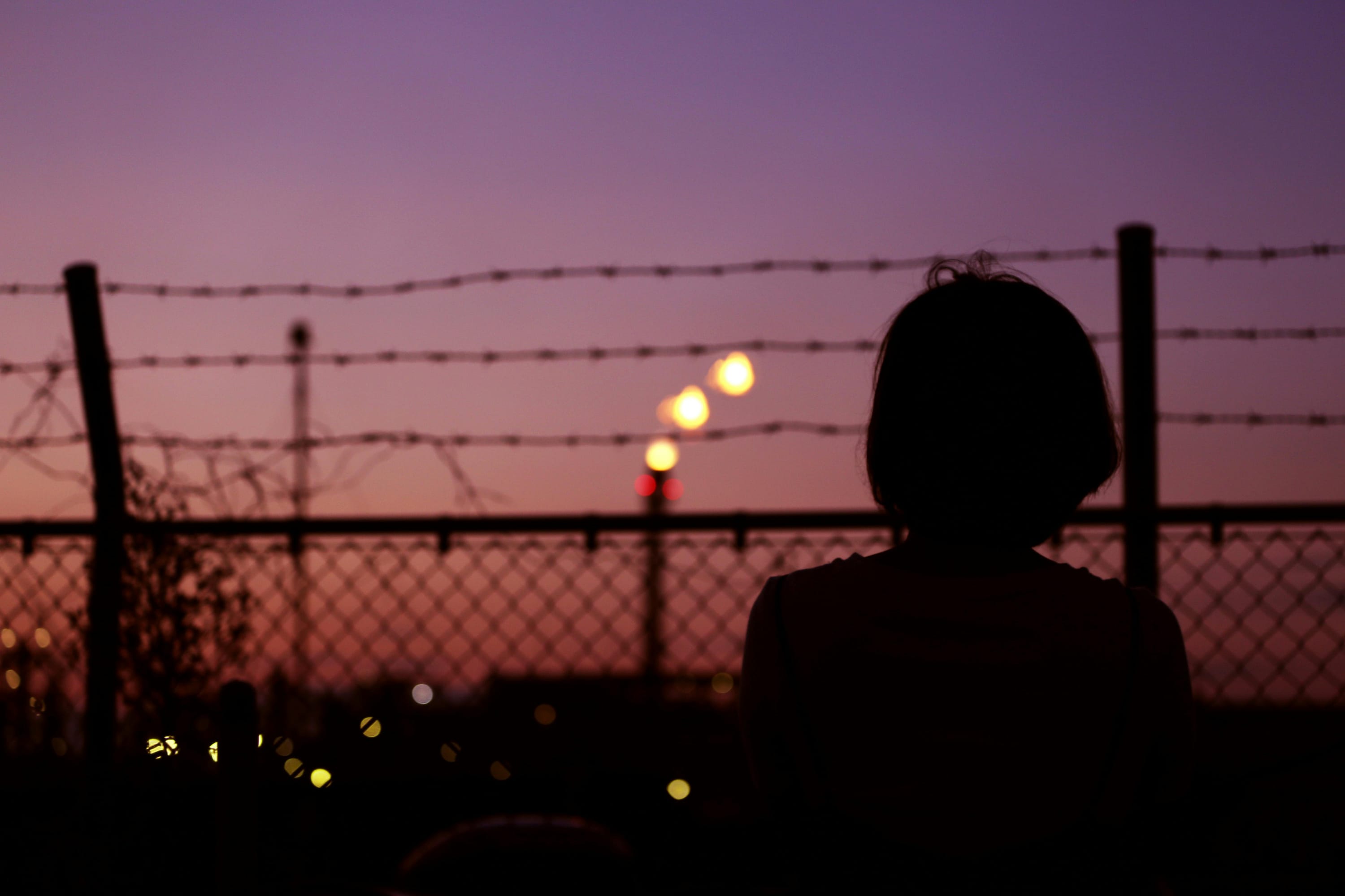 Woman sitting silhouetted looking at the sunset and facing a barbed wire fence